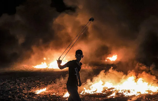 A Palestinian protester swings a slingshot near burning tyres following a demonstration along the border between the Gaza Strip and Israel east of Gaza City on August 28, 2021. (Photo by Mahmud Hams/AFP Photo)