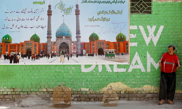 A Hazara man stands beside a poster depicting Jamkaran Mosque in Iran as he keeps an eye on his pair of caged chukars along a street in Mariabad, Quetta, Pakistan, June 19, 2019. (Photo by Akhtar Soomro/Reuters)