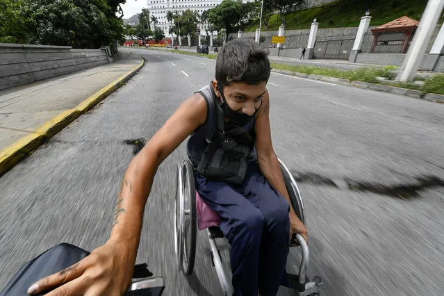 A man wearing a protective face mask hitches a ride in his wheelchair by grabbing on to the back of a motorcycle, near the Miraflores Palace in Caracas, Venezuela, Saturday, August 8, 2020, amid the new coronavirus pandemic. (Photo by Matias Delacroix/AP Photo)