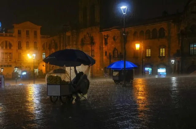 Vendors take cover from a rain shower, as they sit parked outside the San Francisco Basilica, in La Paz, Bolivia, Thursday, January 25, 2024. (Photo by Juan Karita/AP Photo)
