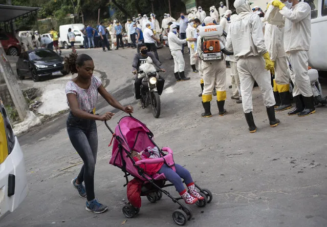 A woman pushing a baby stroller walks past water utility workers from CEDAE who will disinfect the Turano favela in an effort to curb the spread of the new coronavirus in Rio de Janeiro, Tuesday, June 9, 2020. (Photo by Silvia Izquierdo/AP Photo)