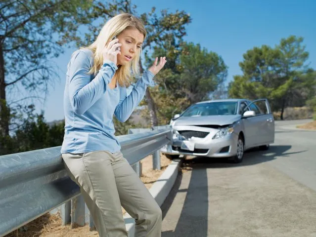 Frustrated woman using cell phone next to car wrecked on guardrail. (Photo by Chris Ryan/Getty Images)