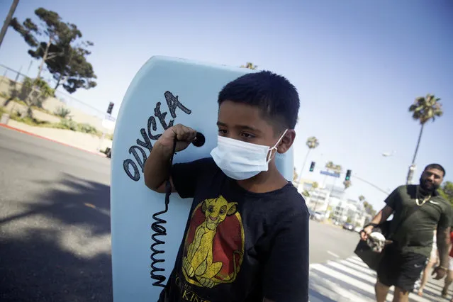 Elijah Cobos wear a face mask as he carries a boogie board to the beach Sunday, July 12, 2020, in Santa Monica, Calif., amid the coronavirus pandemic . A heat wave has brought crowds to California's beaches as the state grappled with a spike in coronavirus infections and hospitalizations. (Photo by Marcio Jose Sanchez/AP Photo)