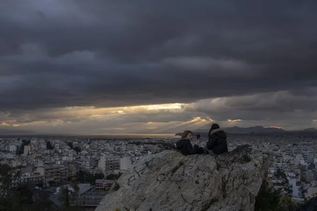 Two young women sit on a rock as sun rays shine through clouds of the Athenian sky while the sun sets behind the Saronic Gulf, on Friday, March 17, 2023. (Photo by Petros Giannakouris/AP Photo)