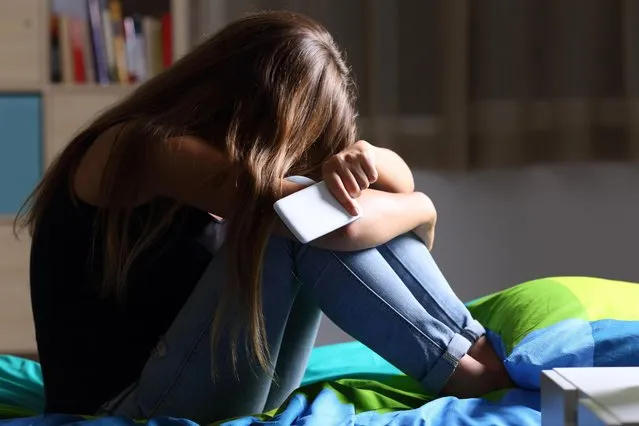 Single sad teen holding a mobile phone lamenting sitting on the bed in her bedroom with a dark light in the background. (Photo by Antonio Guillem/Getty Images)