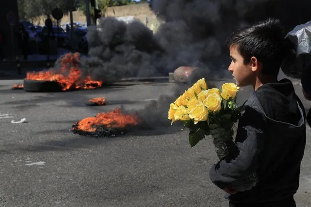 A Syrian boy who sells flowers on the street watches protesters burn tires to block a main road during a protest against the increase in prices of consumer goods and the crash of the local currency, in Beirut, Lebanon, Tuesday, March 16, 2021. (Photo by Hussein Malla/AP Photo)