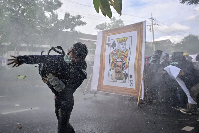 A pro-democracy protester throws an object as others take cover from police water cannon behind a derogatory image of Thailand's Prime Minister Prayut-Chan-O-Cha, as they march to Government House to call for his resignation in Bangkok on July 18, 2021. (Photo by Lillian Suwanrumpha/AFP Photo)