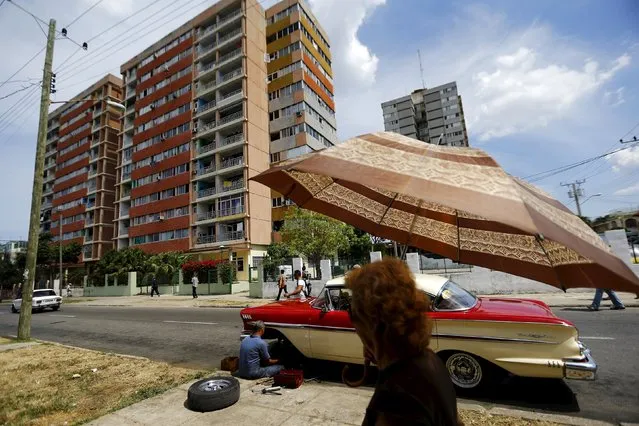 A woman uses an umbrella as a man repairs a vintage car on a street in Havana city, Cuba, March 17, 2016. (Photo by Ivan Alvarado/Reuters)