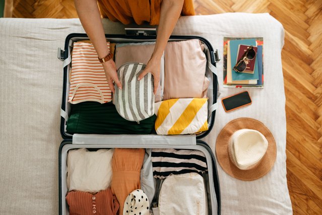 Shot from above of an anonymous woman packing things in her suitcase on the bed. She is holding and putting a grey and white stripped cosmetics bag in. There is her mobile phone, books and sunglasses on the bed. (Photo by miniseries/Getty Images)