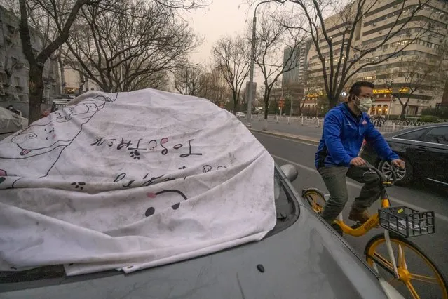 A man wearing a face mask rides past a car with a sheet on the windshield to keep it clean amid dust and haze in Beijing, Friday, March 10, 2023. Skyscrapers disappeared into the haze and air quality plummeted as China's capital was enveloped in a dust storm and heavy pollution on Friday. (Photo by Mark Schiefelbein/AP Photo)