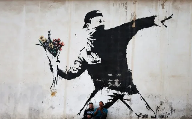 Two men are sitting in front of a famous graffiti of British street artist Banksy, painted on a wall of a gas station in the West Bank city of Bethlehem on December 16, 2015. (Photo by Thomas Coex/AFP Photo)