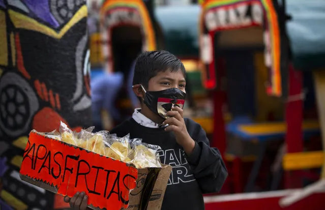 A child, wearing a protective face mask, hawks bags of potato chips near a row of painted wooden boats known as trajineras, popular with tourists that ply the water canals in the Xochimilco district of Mexico City on August 21, 2020, during a reopening of activities after a six-month pause due to the COVID-19 pandemic. (Photo by Marco Ugarte/AP Photo)