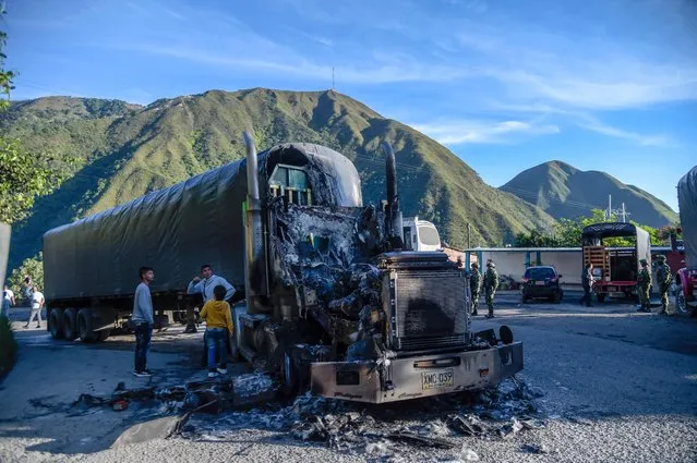 Youths stand next to a burned truck on the Ocaña-Sardinata road, in northeast Colombia, on November 22, 2022. Armed men stopped and burned five trucks on a highway near the border with Venezuela. According to the authorities, the ELN guerrilla and the FARC dissidence operate in that region. The incineration of vehicles occurs with some regularity by illegal armed groups on different roads in Colombia to threaten transporters and the general population and exert territorial control. (Photo by Schneyder Mendoza/AFP Photo)