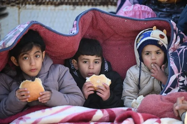 Children eat bread as they sit under a cover in the southeastern Turkish city of Kahramanmaras, on February 8, 2023, two days after a strong earthquake struck the region. Searchers were still pulling survivors on February 8 from the rubble of the earthquake that killed over 11,200 people in Turkey and Syria, even as the window for rescues narrowed. For two days and nights since the 7.8 magnitude quake, thousands of searchers have worked in freezing temperatures to find those still alive under flattened buildings on either side of the border. (Photo by Ozan Kose/AFP Photo)