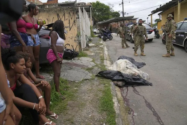 Women watch as military police patrol where the covered body of a person, who was killed during a police operation, lies in the street in the Vila Cruzeiro favela in Rio de Janeiro, Brazil, Friday, February 11, 2022. The police operation against alleged drug traffickers left at least eight people dead. (Photo by Silvia Izquierdo/AP Photo)