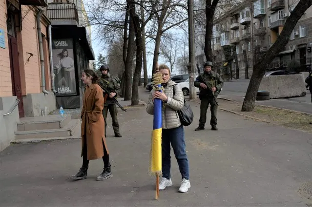 A woman holds a Ukrainian flag near soldiers standing guard outside a government building hit by Russian rockets on March 29, 2022, in Mykolaiv, amid Russian invasion of Ukraine. (Photo by Bulent Kilic/AFP Photo)