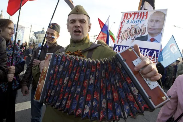 A demonstrator, dressed in Soviet-era military uniform, sings, on his way to a concert marking the one year anniversary of annexation of Ukraine's Crimea peninsula, in downtown Moscow, Russia, Wednesday, March 18, 2015. A poster in the background shows a portrait of Russian President Vladimir Putin and reads “Putin Sows Kindness in the World”. (Photo by Denis Tyrin/AP Photo)