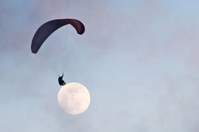 In this photo taken on December 21, 2018, the moon is seen in the distance past a paraglider near Ma On Shan peak in Hong Kong. With most young Hong Kongers priced out of the city's eye-watering property market – often living with parents in cramped flats well into their thirties – paragliding is one of a number of outdoor sports that offers release from the stress of the concrete jungle below. (Photo by Anthony Wallace/AFP Photo)