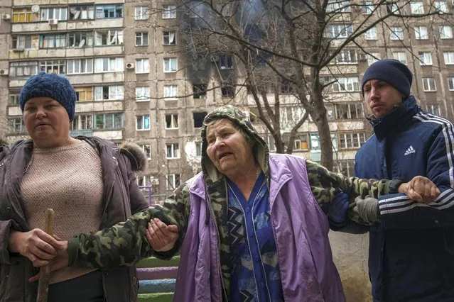 People help an elderly woman to walk in a street with an apartment building hit by shelling in the background in Mariupol, Ukraine, Monday, March 7, 2022. (Photo by Evgeniy Maloletka/AP Photo)
