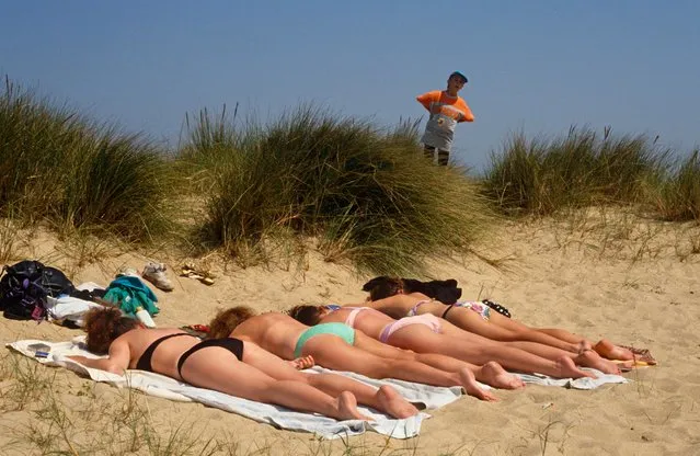A young boy peers over a clump of vegetation to spy on four beautiful women who are all lying face-down in a sandy dune near the seaside resort of “Great Yarmouth”, Norfolk, UK on August 20, 1993. The girls are oblivious to the attention he is giving them and he cheekily stands, undiscovered, with hands on hips. The four females are in bathing costumes with their possessions strewn about the sand. It is a hot afternoon in the sand dunes and the girls are in an otherwise secluded, windless spot. They remain unaware they are being watched by a pre-pubescent voyeur. (Photo by In Pictures Ltd./Corbis via Getty Images)