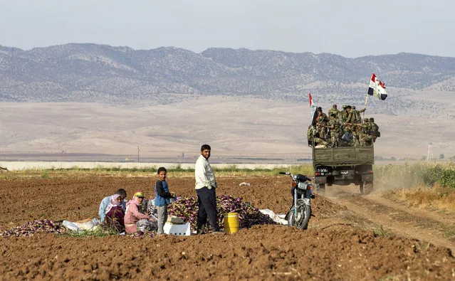 Syrian government soldiers ride in the back of a truck with national flags past people sitting in a field with harvested aubergines, as government forces deploy for the first time in the eastern countryside of the city of Qamishli in the northeastern Hasakah province on November 5, 2019. (Photo by Delil Souleiman/AFP Photo)