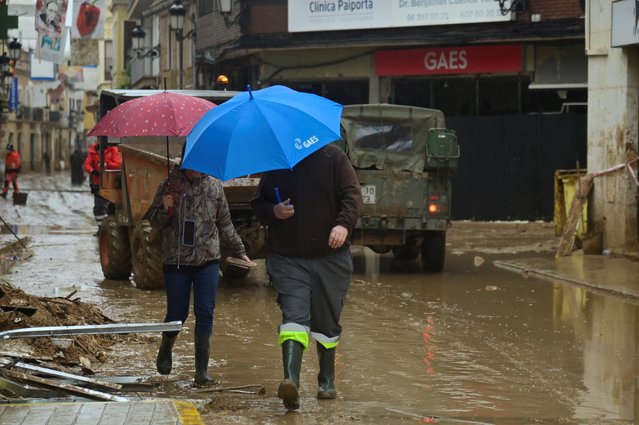 Pedestrians shelter from the rain under umbrellas in a street covered in mud in Paiporta, south of Valencia, eastern Spain, on November 13, 2024 in the aftermath of deadly flooding. Spain closed schools and evacuated residents as heavy rains lashed the country on November 13, two weeks after its worst floods in a generation killed more than 200 people. (Photo by Jose Jordan/AFP Photo)