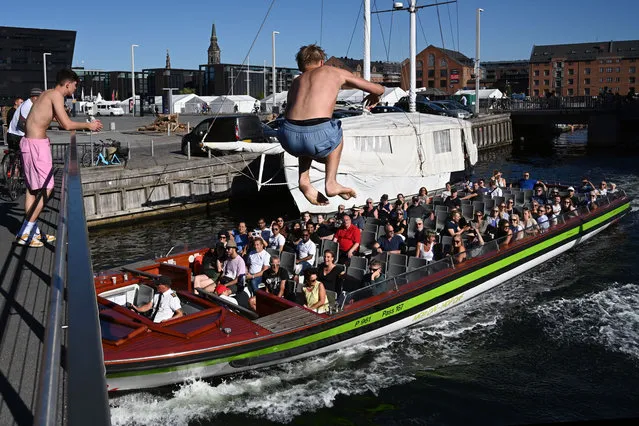 A man jumps from the bridge to cool off tourists in the boat sailing on a canal in Copenhagen, Denmark, on August 24, 2022. On hot days, Copenhageners swim with the city's skyline in free view. During the week, many locals pass the pool for a quick dip on their way home from work. Harbour baths are very popular among both locals and visitors. (Photo by Sergei Gapon/Anadolu Agency via Getty Images)