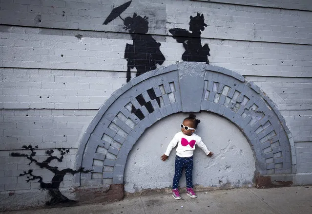 A child poses for a photo under a new art piece by British graffiti artist Banksy in the Brooklyn borough of New York, October 17, 2013. (Photo by Carlo Allegri/Reuters)