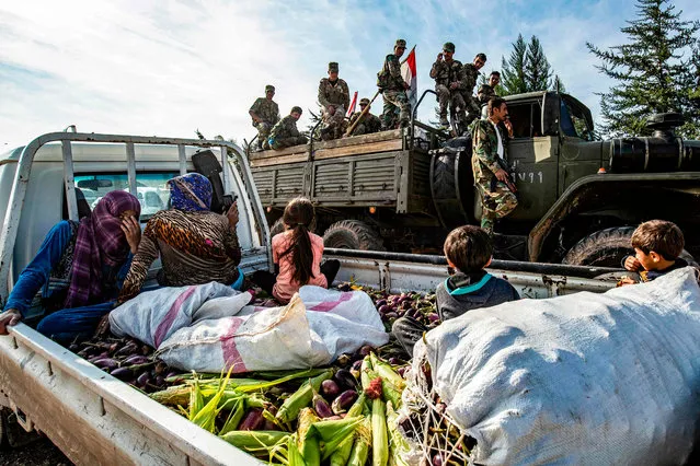 Syrian government soldiers ride in the back of a truck with national flags past a pickup truck carrying women and children seated above harvested aubergines and maize, as government forces deploy for the first time in the eastern countryside of the city of Qamishli in the northeastern Hasakah province on November 5, 2019. (Photo by Delil Souleiman/AFP Photo)