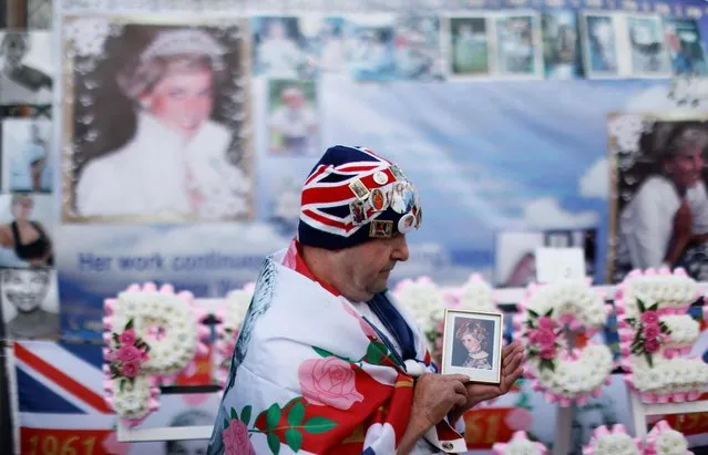 A man wrapped in the British national flag and wearing a hat covered with pins of the British royal family holds a photo portrait of Britain's Princess Diana during a gathering in front of Kensington Palace, in central London, on August 31, 2022 on the 25th anniversary of the Princess of Wales's death. Flowers and other tributes were laid on August 31, 2022 at princess Diana's former London home and above the Paris road tunnel where she lost her life, to mark the 25th anniversary of her death. The former Lady Diana Spencer, whose fairytale marriage to Prince Charles captivated the world until it publicly unravelled with infidelity and divorce, died in a car crash in the French capital on August 31, 1997. (Photo by Carlos Jasso/AFP Photo)