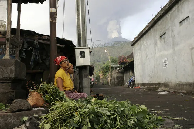 Vegetables sellers wait for customers at a market with a backdrop of the Mount Agung volcano erupting in Karangasem, Indonesia, Monday, November 27, 2017. Indonesia authorities raised the alert for the rumbling volcano to highest level on Monday and closed the international airport on tourist island of Bali stranding thousands of travelers. (Photo by Firdia Lisnawati/AP Photo)