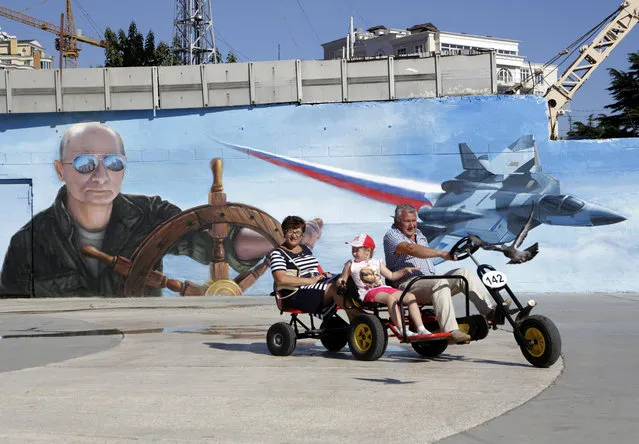 A family rides a quadracycle past a mural depicting Russian President Vladimir Putin in the Crinean city of Yalta, on September 19, 2015. Ukraine has launched a criminal probe into wine-tasting by Russian President Vladimir Putin and ex-Italian premier Silvio Berlusconi after they allegedly quaffed a $100,000 bottle of centuries-old wine in Crimea, prosecutors said. The Kremlin strongman and the disgraced billionaire reportedly cracked open a bottle of 1775 Spanish Jerez de la Frontera at the famed Massandra winery during a private visit by Berlusconi this month to the region Moscow annexed from Kiev in 2014. (Photo by Max Vetrov/AFP Photo)
