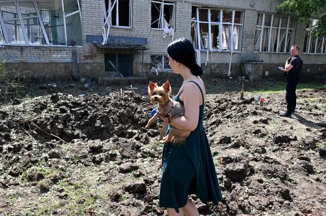 A woman holding a dog stands by a huge crater made from the hit of a rocket close to the destroyed school building in Kharkiv, on July 4, 2022, amid Russian invasion of Ukraine. (Photo by Sergey Bobok/AFP Photo)