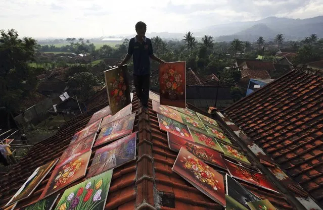 An artist walks across the roof of his house while carrying paintings to be dried at Jelekong village near Bandung, in Indonesia's West Java province, March 20, 2014. The people of Jelekong village earn their living by selling their paintings. Around 300 painters live in the village, producing about 1,000 paintings a month, each priced between 50,000 rupiah to 11,000,000 rupiah ($4.50 to $1000), according to villager Asep Sancang, who works as a high school art teacher and painter. (Photo by Reuters/Beawiharta)
