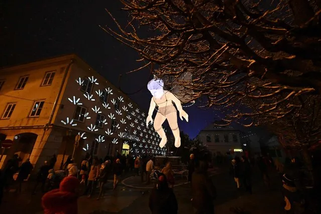 Tourists walk below an inflatable and illuminated puppet that is seen hanging over a street in Veszprem, Hungary on January 21, 2023, as the town and its region of Bakony-Balaton celebrate their title of the 2023 European Capital of Culture. (Photo by Attila Kisbenedek/AFP Photo)