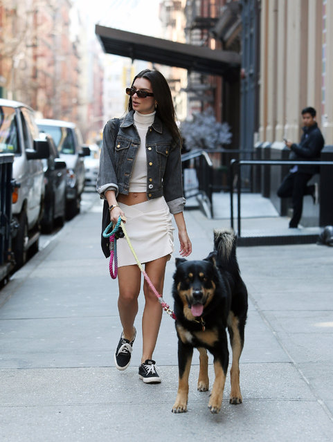 Emily Ratajkowski is pictured walking her dog in New York City on March 16, 2022. The model and author wore a denim jacket, cropped turtleneck, white mini skirt, and black canvas trainers. (Photo by The Image Direct)