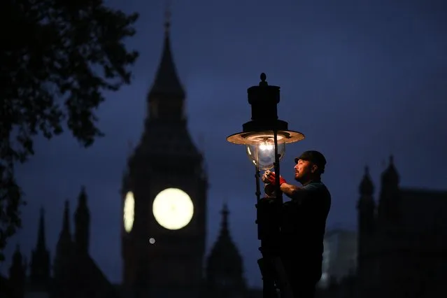 A British Gas engineer services a gas-powered lamp near the Houses of Parliament in central london on November 11, 2022. Intrigued tourists watch as Paul Doy climbs a ladder outside London's Westminster Abbey and lifts the globe of a gas street lamp. Winding its timer, he then ignites a small cloth mesh, creating a distinctive soft warm light that illuminates the darkness. “I like the historical aspect of it”, said Doy, even if it means getting up at 5:00 am to tend to the lamps in the fashionable district of Covent Garden. (Photo by Daniel Leal/AFP Photo)