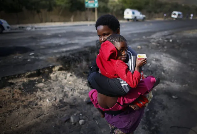 A South African woman carries two sick children to hospital past a barricade during a violent protest of thousands of residents against the police in Masiphumelele, Cape Town, South Africa 23 October 2015. Community members are angered at the arrest of some residents suspected to be vigilantes following the killing of suspected criminals by vigilantes. This follows weeks of protests over the lack of policing in the impoverished area and the alleged targetting of vigilante members by the police. (Photo by Nic Bothma/EPA)