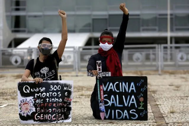Demonstrators protest the president's handling of the COVID-19 pandemic outside Planalto presidential palace in Brasilia, Brazil, Friday, March 19, 2021. The signs read in Portuguese “Almost 300,000 dead. Bolsonaro genocide”, left, and “Vaccinations save lives”. (Photo by Eraldo Peres/AP Photo)