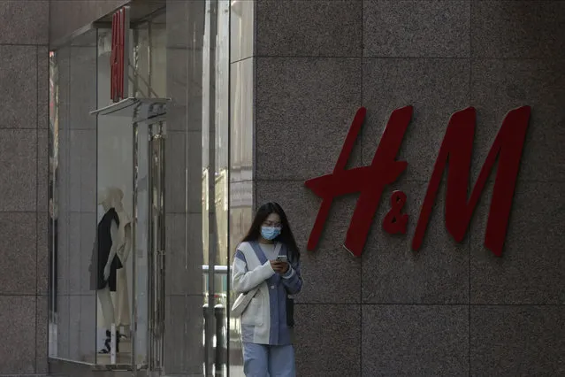 A woman wearing a face mask browses her smartphone as she passes by a Swedish fashion brand H&M store outlet in Beijing, Thursday, March 25, 2021. (Photo by Andy Wong/AP Photo)