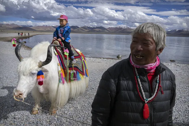 A Tibetan man stands nearby as a young tourist poses for photos on a white yak at the shore of the lake in Namtso in western China’s Tibet Autonomous Region, as seen during a government organized visit for foreign journalists, Wednesday, June 2, 2021. (Photo by Mark Schiefelbein/AP Photo)