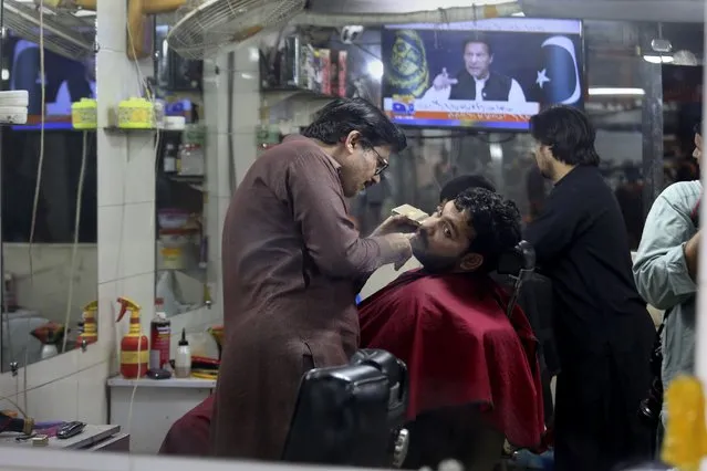 A barber deals a customer as a news channel broadcast a live address to the nation by Pakistan's Prime Minister Imran Khan at a market, in Peshawar, Pakistan, Thursday, March 31, 2022. Pakistan's embattled Prime Minister Khan remained defiant on Thursday, telling the nation that he will not resign even as he faces a no-confidence vote in parliament and the country's opposition says it has the numbers to push him out. (Photo by Muhammad Sajjad/AP Photo)