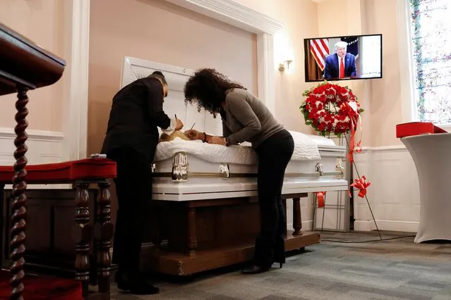 Jenny Adames, 36, a Funeral Service Coordinator at International Funeral & Cremation Services, a funeral home in Harlem, applies makeup to her aunt, a suspected COVID-19 victim, with the assistance of her cousin Vanessa Fernandez, before her aunt's viewing service, as a screen displays a media conference by U.S. President Donald Trump, during the coronavirus disease (COVID-19) outbreak, in Manhattan, New York City, New York, U.S., April 14, 2020. (Photo by Andrew Kelly/Reuters)