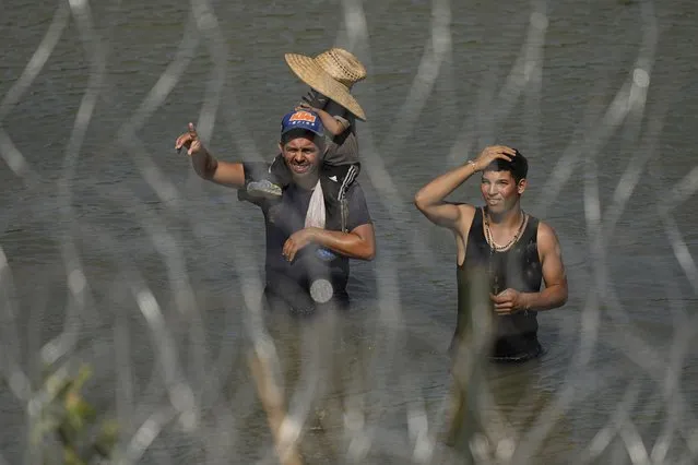 Migrants stand in the Rio Grande behind concertina wire as they try to enter the U.S. from Mexico near the site where workers are assembling large buoys to be used as a border barrier in Eagle Pass, Texas, Tuesday, July 11, 2023. The floating barrier is being deployed in an effort to block migrants from entering Texas from Mexico. (Photo by Eric Gay/AP Photo)