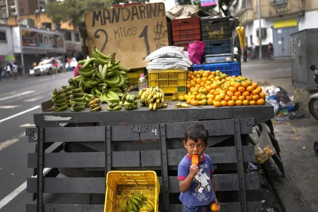 A child eats ice cream near a truck that sells fruit with prices set in US dollars in Caracas, Venezuela, Sunday, December 11, 2022. On Saturday, President Nicolas Maduro announced he had ordered his team to implement measures “in defense” of the official exchange rate that is established by the country's central bank. (Photo by Matias Delacroix/AP Photo)