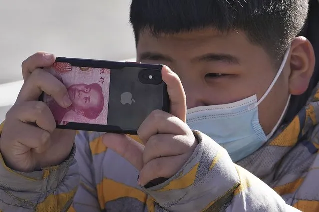 A boy wearing a face mask using an iPhone with a Chinese currency note takes a picture on a street in Beijing, Tuesday, January 17, 2023. China's economic growth fell to its second-lowest level in at least four decades last year under pressure from anti-virus controls and a real estate slump, but activity is reviving after restrictions that kept millions of people at home and sparked protests were lifted. (Photo by Andy Wong/AP Photo)