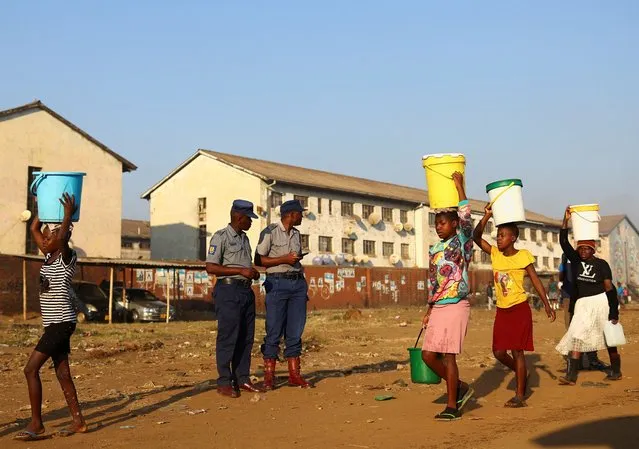 Police officers look on as women walk past carrying buckets filled with water ahead of the presidential elections in Mbare township, in Zimbabwe on August 22, 2023. (Photo by Siphiwe Sibeko/Reuters)
