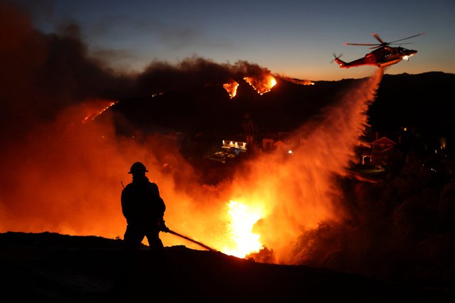 Fire personnel respond to homes destroyed while a helicopter drops water as the Palisades Fire grows in Pacific Palisades, California on January 7, 2025. A fast-moving wildfire in a Los Angeles suburb burned buildings and sparked panic, with thousands ordered to evacuate January 7, 2025 as "life threatening" winds whipped the region. Frightened residents abandoned their cars on one of the only roads in and out of the upscale Pacific Palisades area, fleeing on foot from the 770-acre (310-hectare) blaze engulfing an area crammed with multi-million dollar homes in the Santa Monica Mountains. (Photo by David Swanson/AFP Photo)