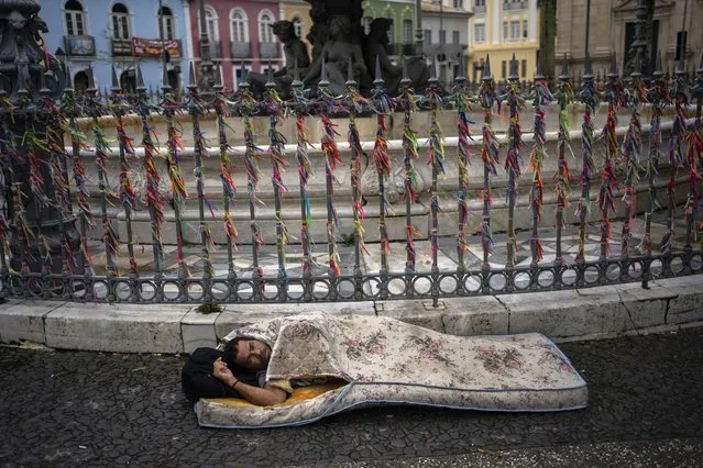 Homeless Joao De Jesus rests inside a mattress on a rainy day in the Pelourinho neighborhood of Salvador, Bahia state, Brazil, early Monday, September 19, 2022. Behind De Jesus are “Bonfim” ribbons tied to a fence, which according to popular belief are synonymous with faith and tradition and have the power to make three wishes come true. (Photo by Rodrigo Abd/AP Photo)