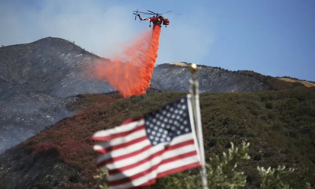 Framed by a burned-over hillside, rear, and an American flag in the foreground, a helicopter drops fire retardant ahead of advancing flames as a wildfire approaches Placerita Canyon in Santa Clarita, Calif., Monday, July 25, 2016. (Photo by Nick Ut/AP Photo)